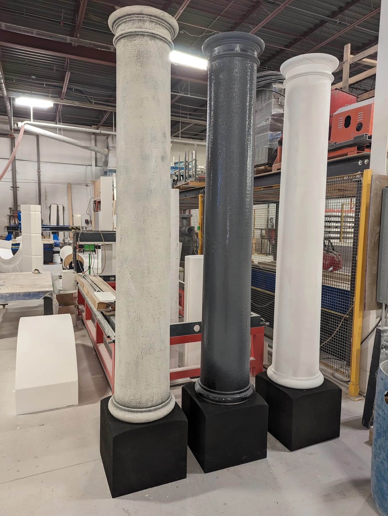 Three tall decorative columns on black plinth bases in the workshop showing stone-textured, dark polyurea, and smooth white finishes.