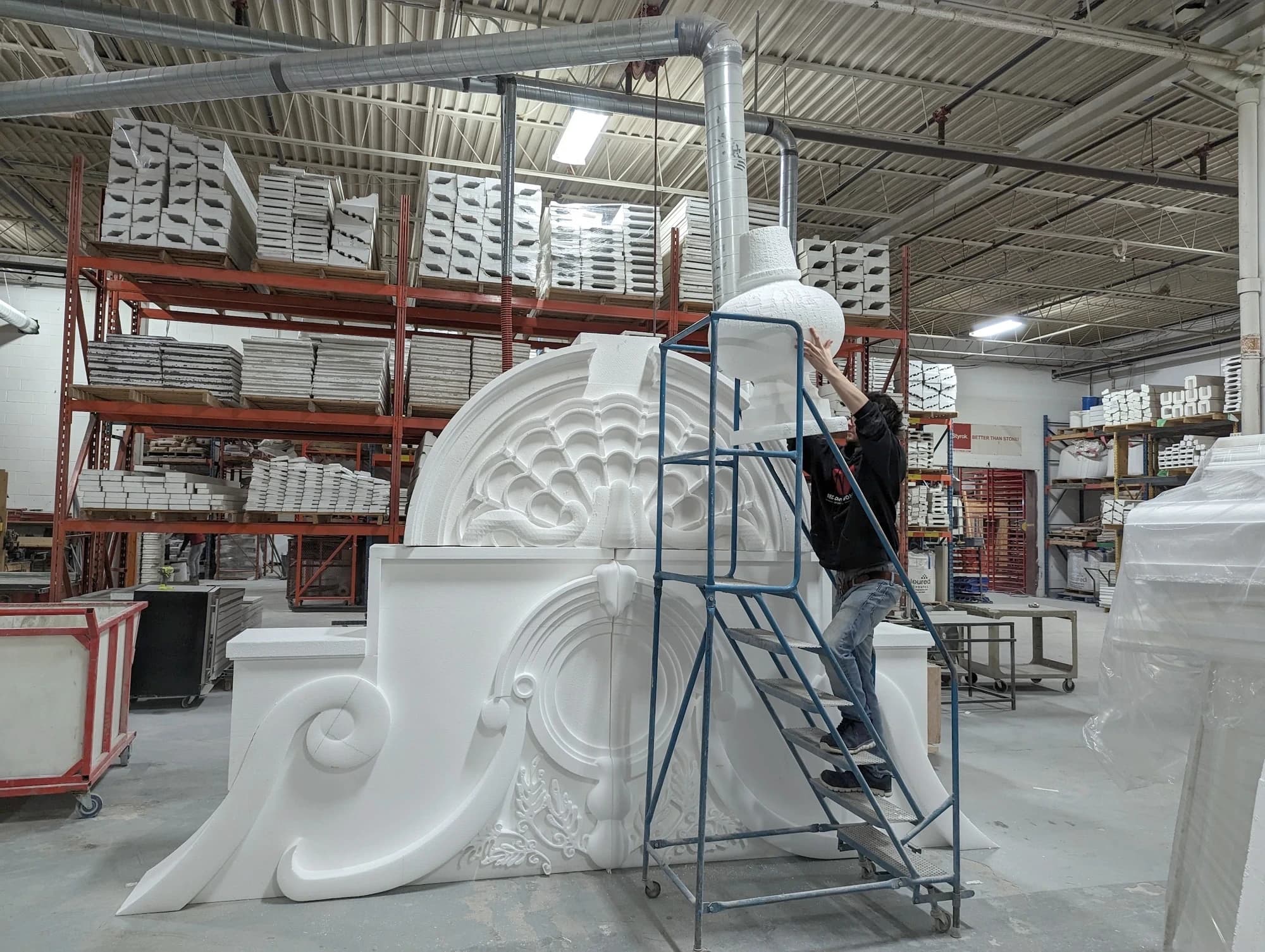 Large ornate white foam assembly with scrollwork, curved mouldings, and a central shell motif beside a rolling workshop ladder.
